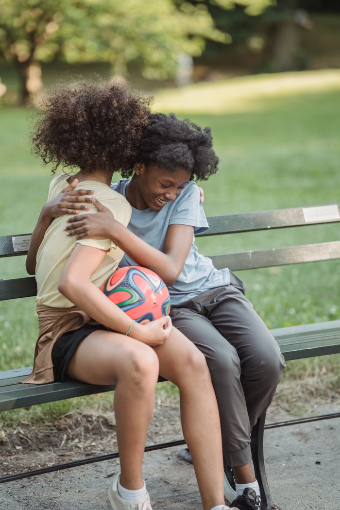 creative-03 Two friends sharing a joyful hug while sitting on a park bench with a soccer ball.
