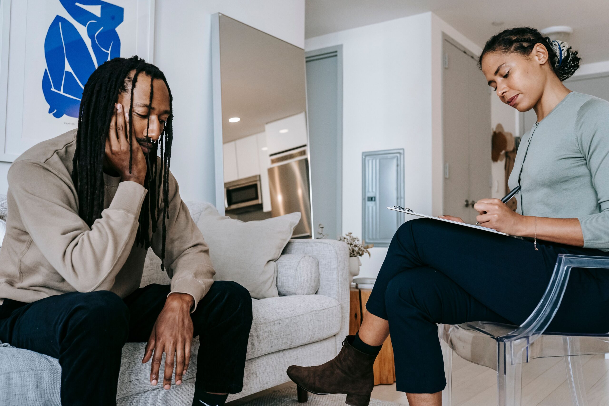 Attentive African American female with clipboard taking notes near sad male partner touching face on couch at home